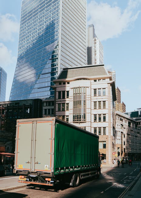 A large green and white box truck parked on a city street in West Hampstead, with tall modern glass skyscrapers and historic stone buildings in the background. The truck is positioned close to the sidewalk, with its rear doors closed, ready for loading or unloading furniture and packing materials. The scene is captured during daytime under clear blue skies, with pedestrians and cyclists visible nearby. This is a typical setting for a house or office removal, where furniture and boxes are transported in a vehicle as part of a home relocation or professional moving service. The image reflects the urban environment involved in furniture transport and packing and moving logistics, with [COMPANY_NAME] likely engaged in a full-service removals operation as part of the West Hampstead moving process.