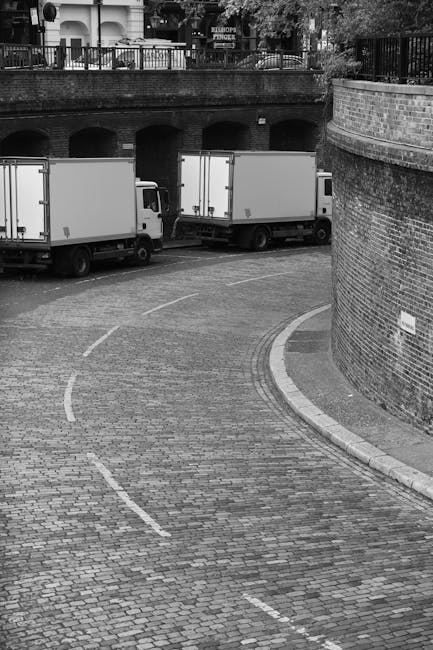 A black-and-white photograph shows a narrow cobbled street with a curved section, bordered by a brick wall on the right side and a metal railing on the left, above which a brick balcony with trees and residential buildings is visible. Parked on the street are two medium-sized box trucks, positioned side by side near the curb, with the side doors closed. The trucks are part of a home relocation or removals process managed by a professional moving company, possibly referred to as [COMPANY_NAME], involved in furniture transport and packing operations. The setting indicates a quiet urban area with limited traffic, and the empty cobblestone surface suggests preparation for loading or unloading furniture, boxes, and packing materials, which are typical elements in house removals. The scene captures a moment of logistical movement, with the trucks poised to facilitate the transport of belongings during a domestic move in West Hampstead, London.