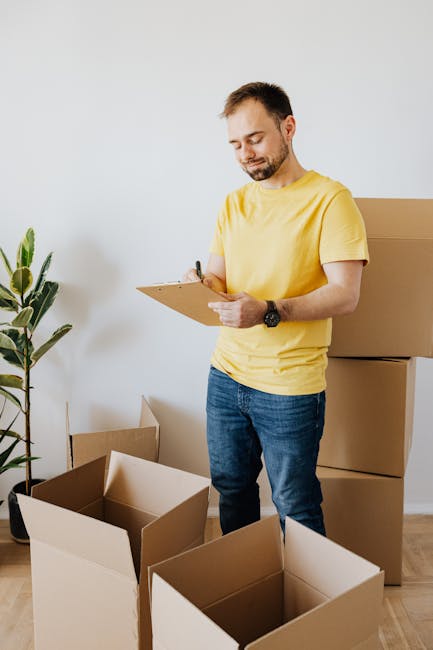 A man wearing a yellow t-shirt and jeans is standing inside a room, holding a clipboard and a pen while reviewing a packing list. Surrounding him are several open cardboard boxes, some filled with packing materials such as paper and plastic wrap. Behind him, additional sealed and unsealed boxes are stacked against a white wall, and a green potted plant is visible in the corner. The scene appears to depict the packing stage of a home relocation, with the individual likely organizing or preparing items for transport. The environment is well-lit with natural light, and the setting suggests an ongoing furniture transport and packing process typical of house removals, as coordinated by companies like [COMPANY_NAME] for West Hampstead moves in NW6.