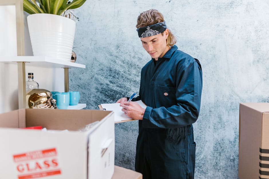 A young man with shoulder-length blond hair, wearing a black bandana and a dark blue work jumpsuit, is inside a room during a house removal process. He is holding a blue pen and a clipboard, appearing to check or record inventory. In front of him, there is a cardboard box with red and white shipping labels, partially open, filled with various household items. The background includes a light, textured wall and white shelving units holding decorative items such as a large white pot with a plant, a glass decanter, and teal cups. The scene takes place in a well-lit interior space, possibly a kitchen or storage area, where the man is preparing for the furniture transport and packing stage of relocation. The setting indicates careful organization and documentation as part of the packing and moving process, supported by [COMPANY_NAME], specializing in residential removals and home relocation services, emphasizing professional logistics and furniture transport.