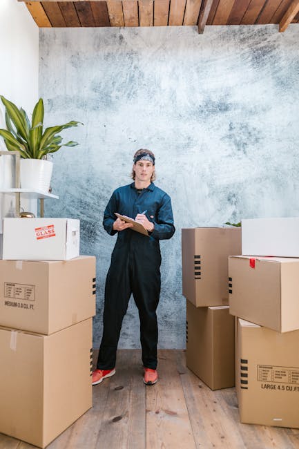 A man wearing a yellow t-shirt and jeans is standing inside a room, holding a clipboard and a pen while reviewing a packing list. Surrounding him are several open cardboard boxes, some filled with packing materials such as paper and plastic wrap. Behind him, additional sealed and unsealed boxes are stacked against a white wall, and a green potted plant is visible in the corner. The scene appears to depict the packing stage of a home relocation, with the individual likely organizing or preparing items for transport. The environment is well-lit with natural light, and the setting suggests an ongoing furniture transport and packing process typical of house removals, as coordinated by companies like [COMPANY_NAME] for West Hampstead moves in NW6.