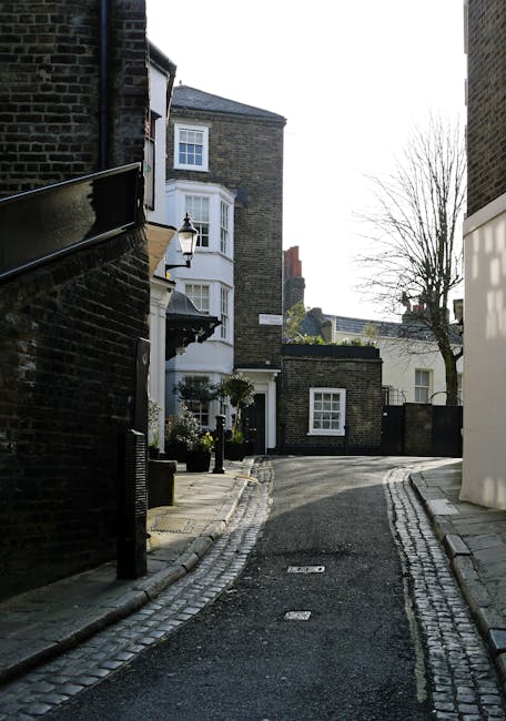 A narrow residential street in West Hampstead, with cobbled pavement and a slight curve, lined with historic brick houses featuring bay windows and small front gardens. On the left, a black brick building has a black metal railing along the sidewalk, with a traditional lamppost mounted on the front wall. In the background, a white building with large windows and a sloped roof is visible, while to the right, there is a modern structure with smooth white paneling. Bare trees are seen on the right side, indicating a winter season. The scene is well-lit by natural daylight, with shadows cast on the pavement and building facades, creating a quiet, characteristic setting for house removals or home relocation activities in the area, possibly involving furniture transport or the unloading of moving boxes contracted through [COMPANY_NAME].