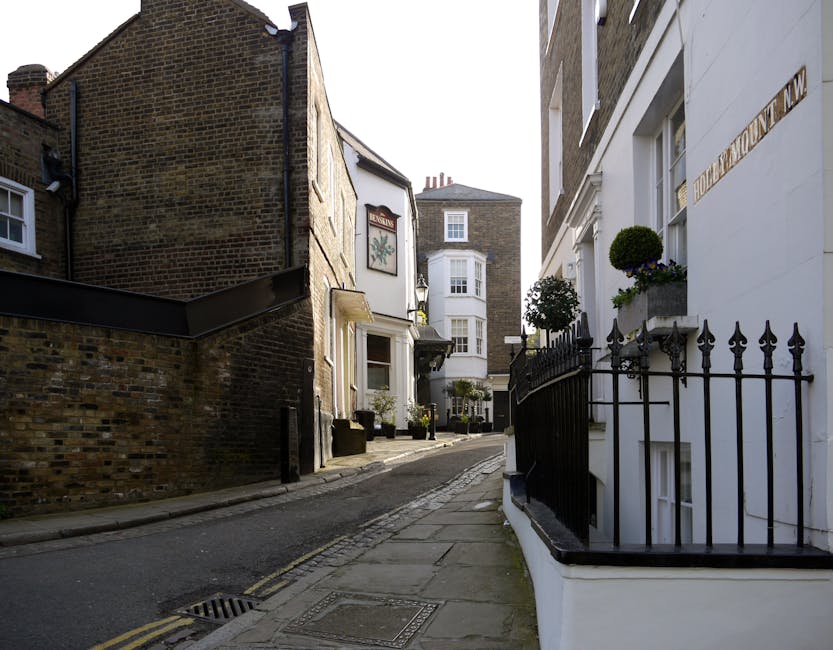 A narrow residential street in West Hampstead showing a mix of historic brick and white-painted buildings with decorative elements. The street is paved with cobblestones and features a gentle curve, with a slight incline. On the right side, there is a white building with black wrought iron railings, flower boxes with small green shrubs, and large ground-floor windows. The left side has a brick building with a painted sign above a doorway and a small bay window. Several large cardboard boxes, wrapped in plastic, are stacked outside near the white building, suggesting packing in preparation for home relocation. A black trolley or dolly is positioned nearby for furniture transport, while the pavement is mostly clear, indicating an organized moving process. Dappled sunlight illuminates the scene, highlighting the details of the buildings and street, capturing a typical street view involved in local removals and house moving services, as would be coordinated by [COMPANY_NAME] in the West End Lane area, supporting furniture transport, packing, and the loading process during house removals.