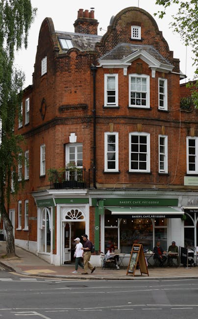 A narrow residential street in West Hampstead, with cobbled pavement and a slight curve, lined with historic brick houses featuring bay windows and small front gardens. On the left, a black brick building has a black metal railing along the sidewalk, with a traditional lamppost mounted on the front wall. In the background, a white building with large windows and a sloped roof is visible, while to the right, there is a modern structure with smooth white paneling. Bare trees are seen on the right side, indicating a winter season. The scene is well-lit by natural daylight, with shadows cast on the pavement and building facades, creating a quiet, characteristic setting for house removals or home relocation activities in the area, possibly involving furniture transport or the unloading of moving boxes contracted through [COMPANY_NAME].